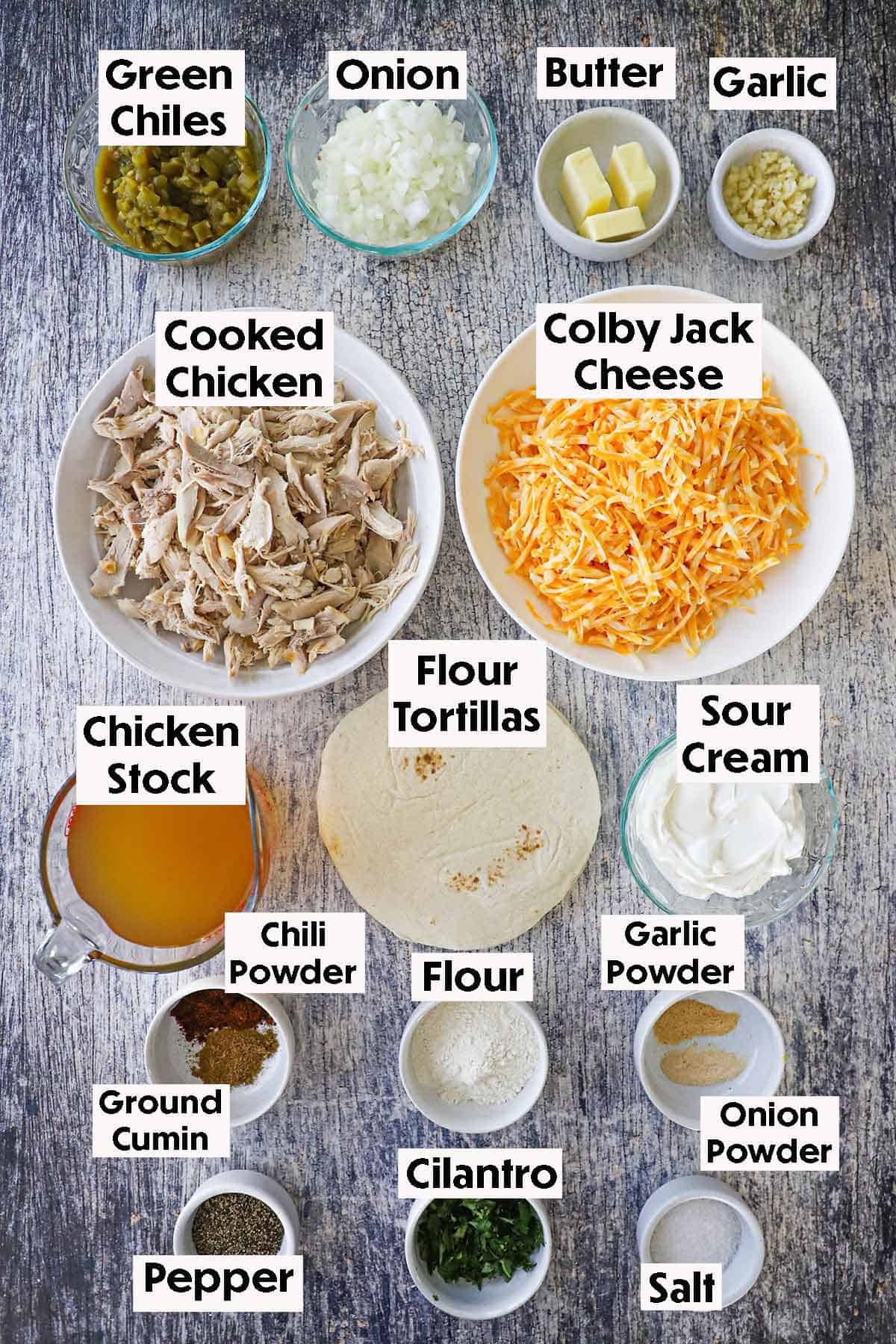 An arrangement of ingredients for chicken enchiladas with sour cream sauce on a grey wooden background including cooked shredded chicken, shredded cheese, chiles, onions, garlic, chicken stock, tortillas, sour cream, and seasonings.