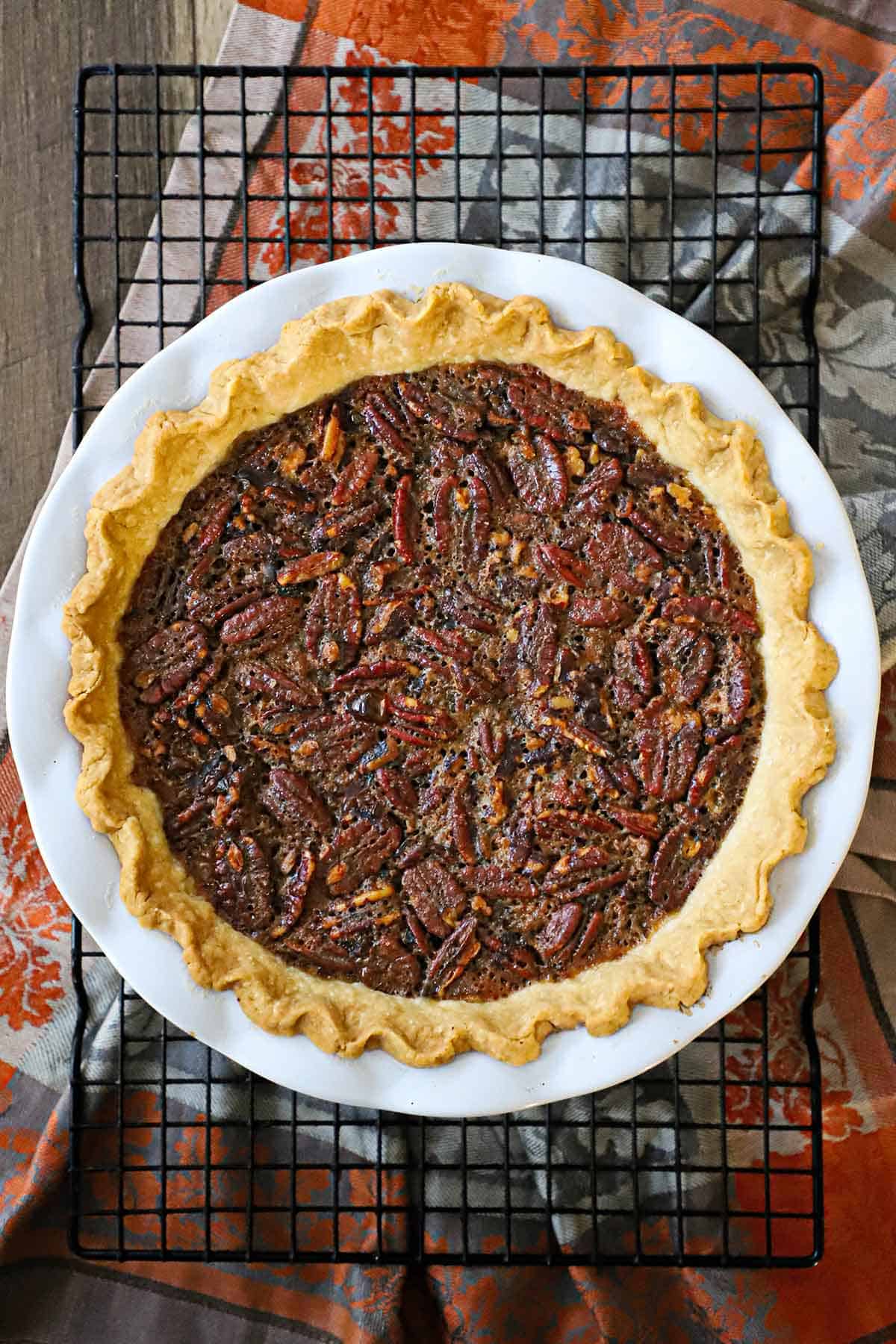 An overhead view of the best chocolate bourbon pecan pie in a white pie dish resting on a baking rack on an autumnal linen underneath.