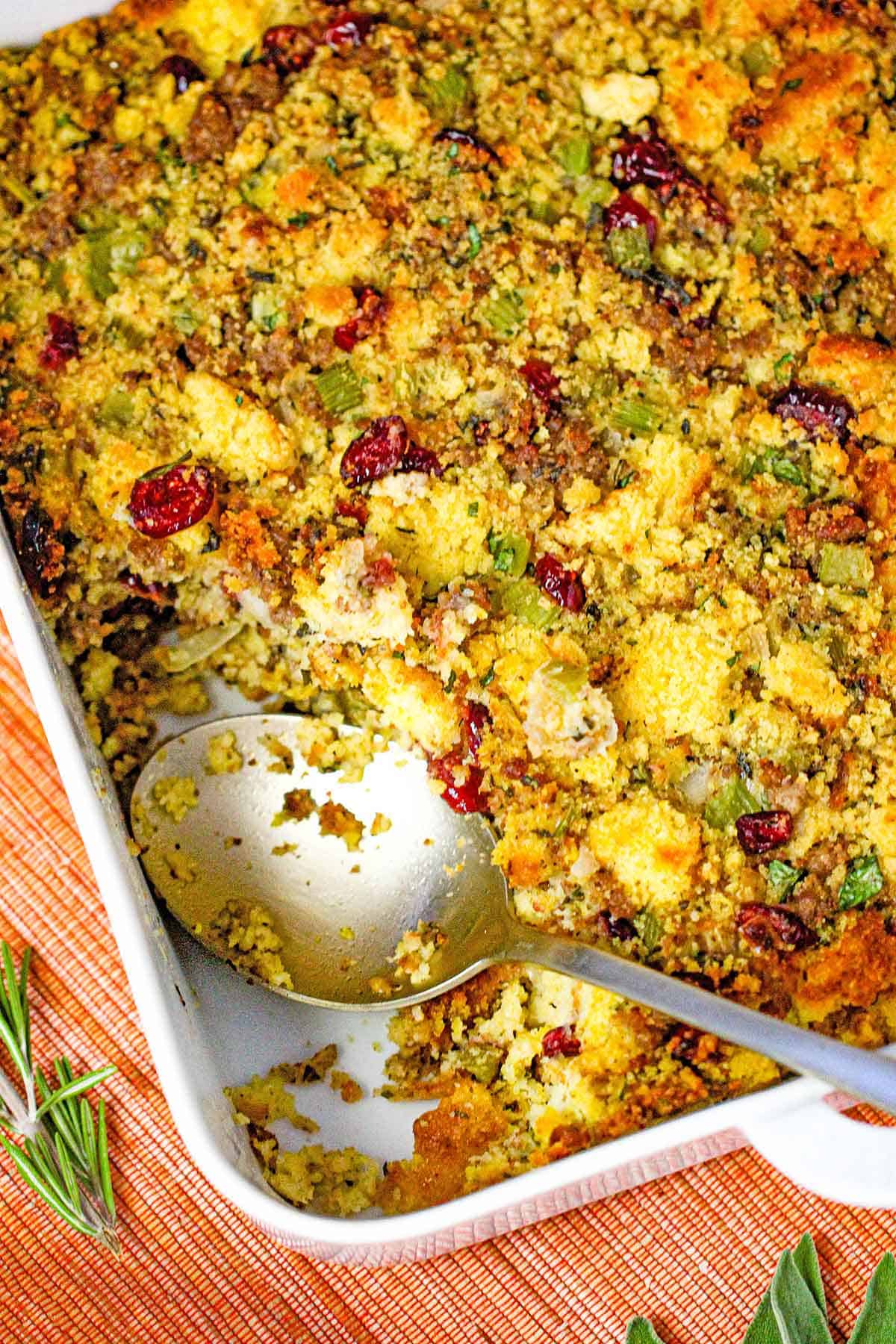 An overhead view of a square white baking dish filled with cornbread dressing with sausage and cranberries with a portion missing with a large spoon inserted into the pan.