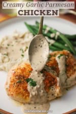 A person using a spoon to drizzle a garlic Parmesan sauce over the top of a seared boneless chicken breast on a white dinner plate.