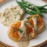 A close-up view of two filets of creamy garlic parmesan chicken on a white dinner plate next a pile of mashed potatoes next to a serving of steamed string beans.