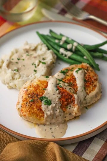 A white dinner plate filled with a serving of creamy garlic parmesan chicken with sauce drizzled over the tops of the filets resting next to a pile of mashed potatoes and steamed string beans.