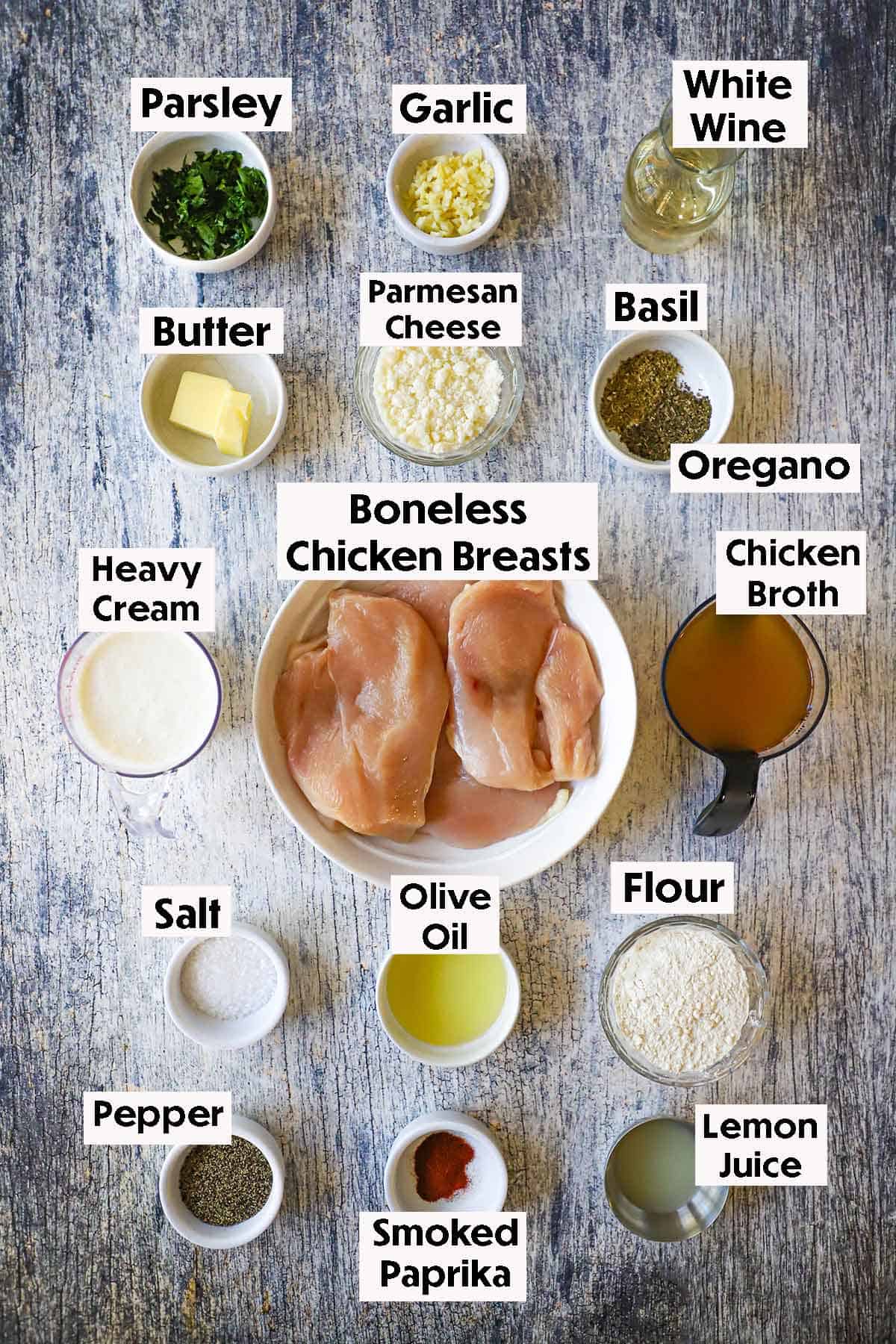 An arrangement of ingredients for creamy garlic Parmesan chicken on a grey wooden background including uncooked boneless chicken breasts, garlic, wine, heavy cream, chicken broth, flour, lemon juice, and seasonings.