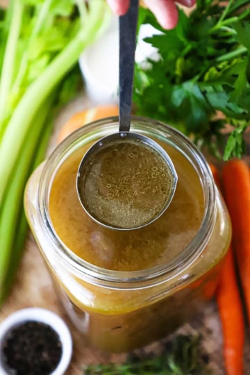 An overhead view of a large ladle of homemade roasted turkey stock being held over a large jar filled with more turkey stock.