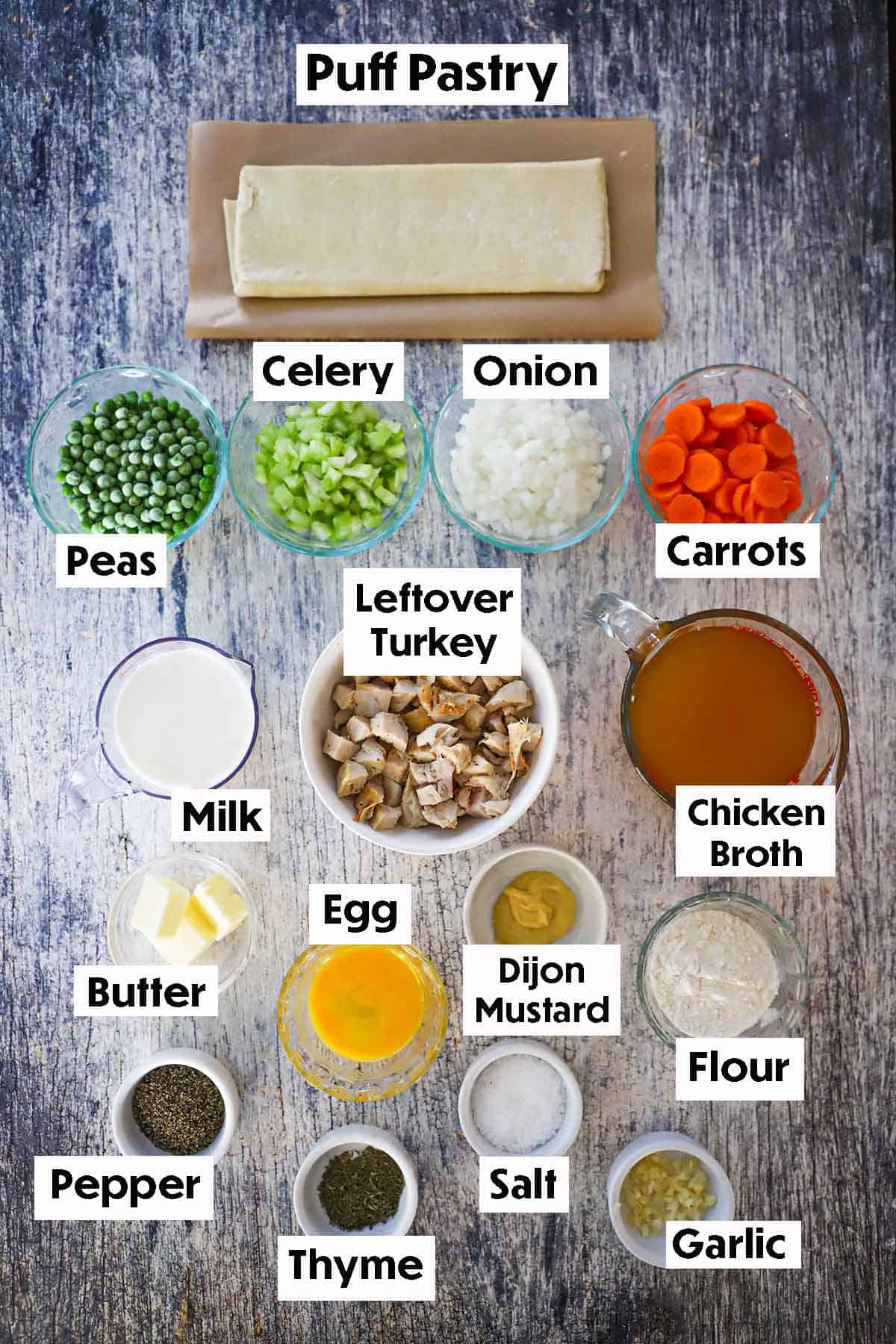 An arrangement of ingredients for Leftover Turkey Pot Pie with Buttery Puff Pastry on a grey wooden background including leftover cubed turkey, celery, onion, carrots, peas, milk, broth, butter, egg, mustard, flour seasonings, and a puff pastry.