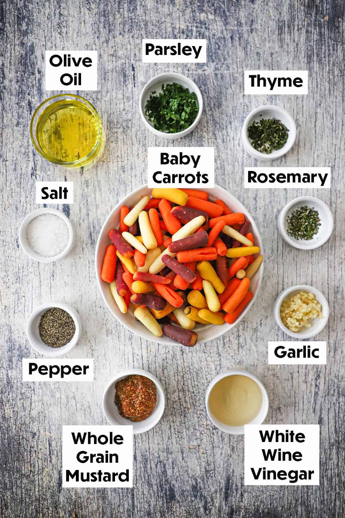 An arrangement of ingredients for roasted baby carrots with herb vinaigrette on a grey wooden background including yellow, orange, and purple baby carrots, oil, whole grain mustard, vinegar, garlic, herbs, and spices.