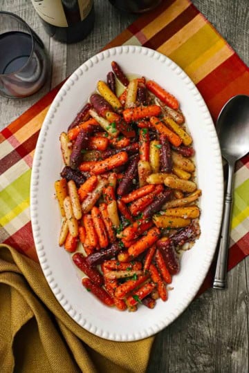 An overhead view of an oval white platter filled with roasted baby carrots with herbed vinaigrette on an autumnal-style place matt.