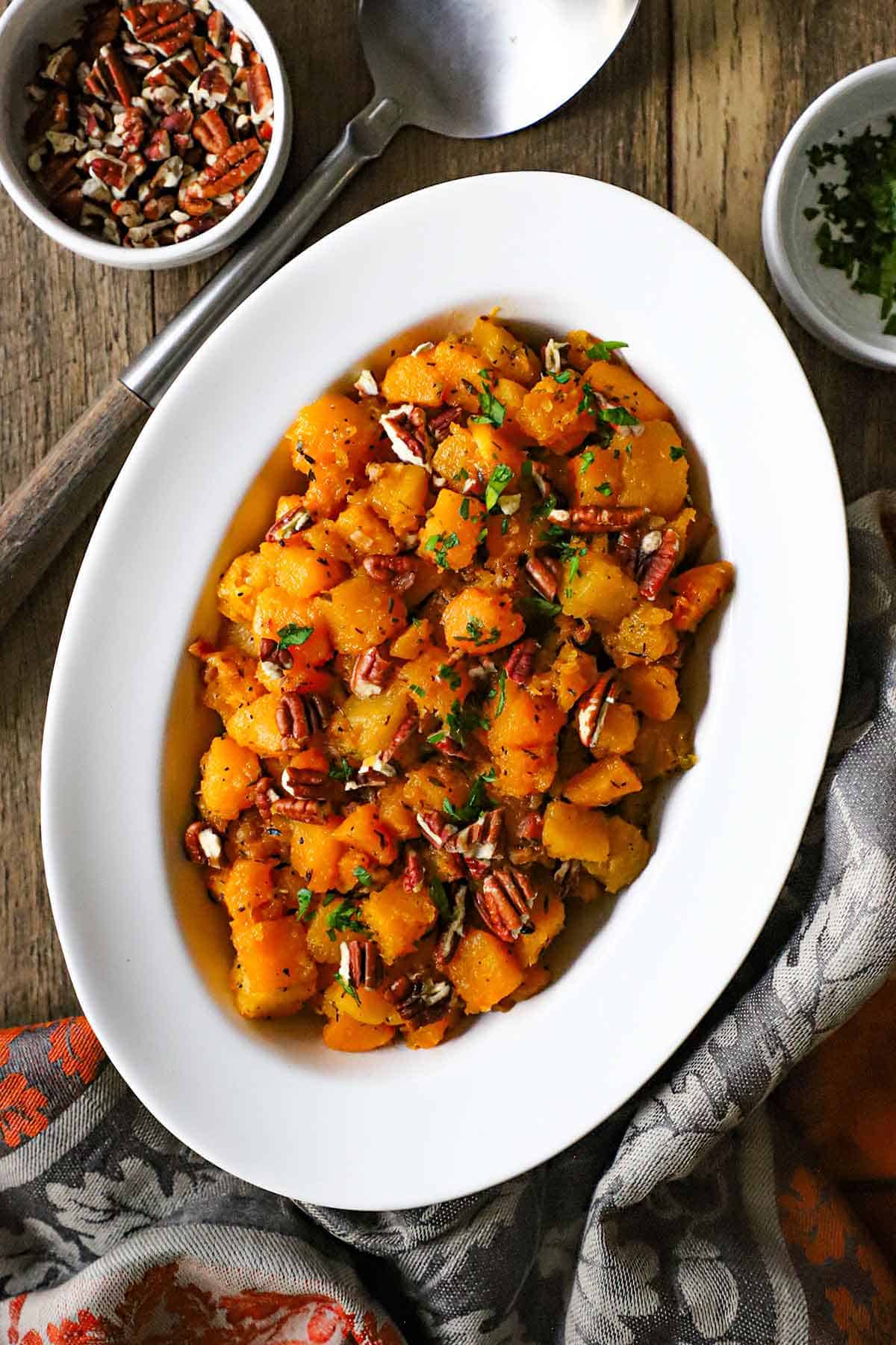 An overhead view of a white oval serving dish filled with Maple-Glazed Butternut Squash with Pancetta and Herbs sitting next to a small bowl of pecans and another bowl of chopped fresh herbs.
