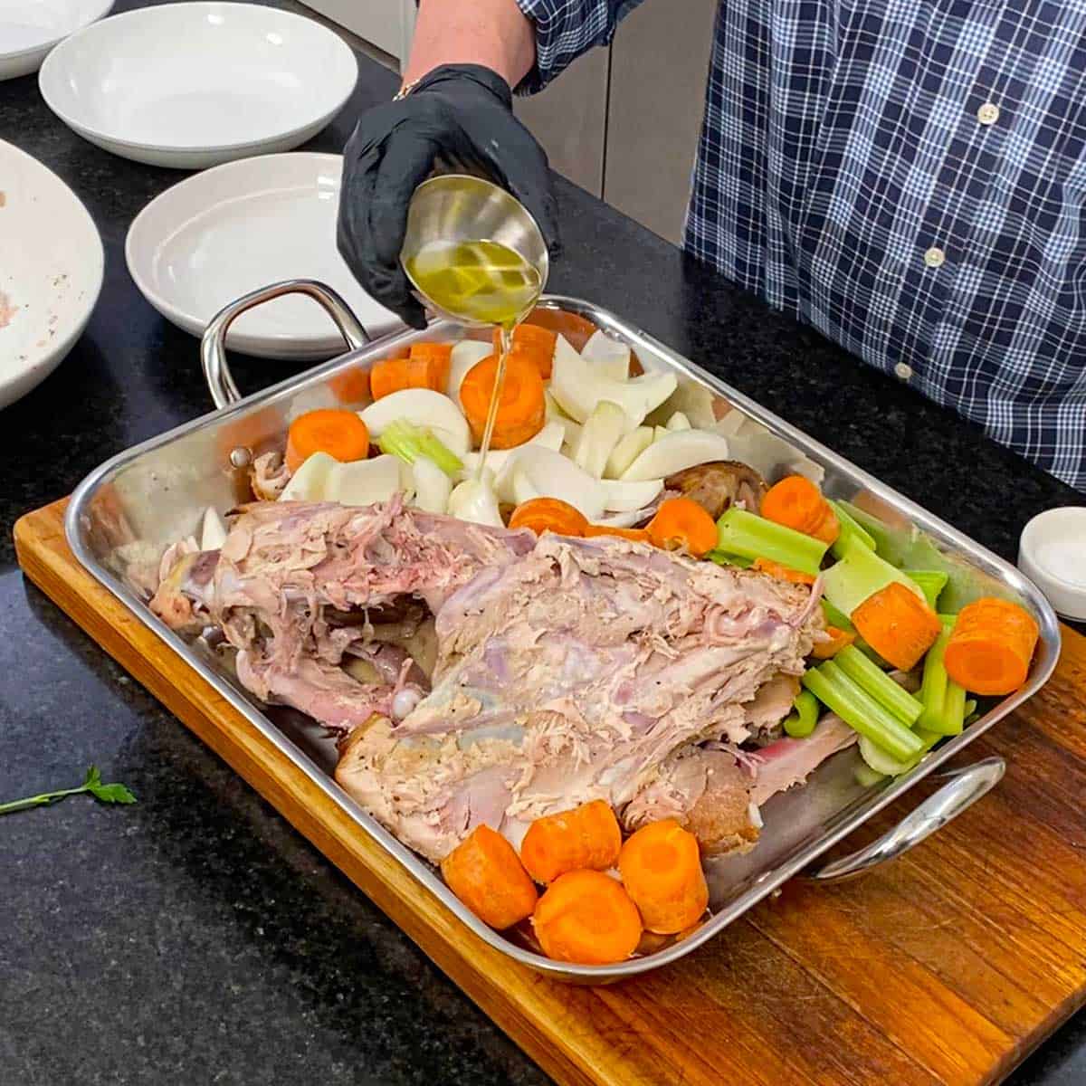 A person pouring olive oil from a small silver bowl over a large roasting pan filled with a turkey carcass and roughly chopped carrots, celery, and onions.