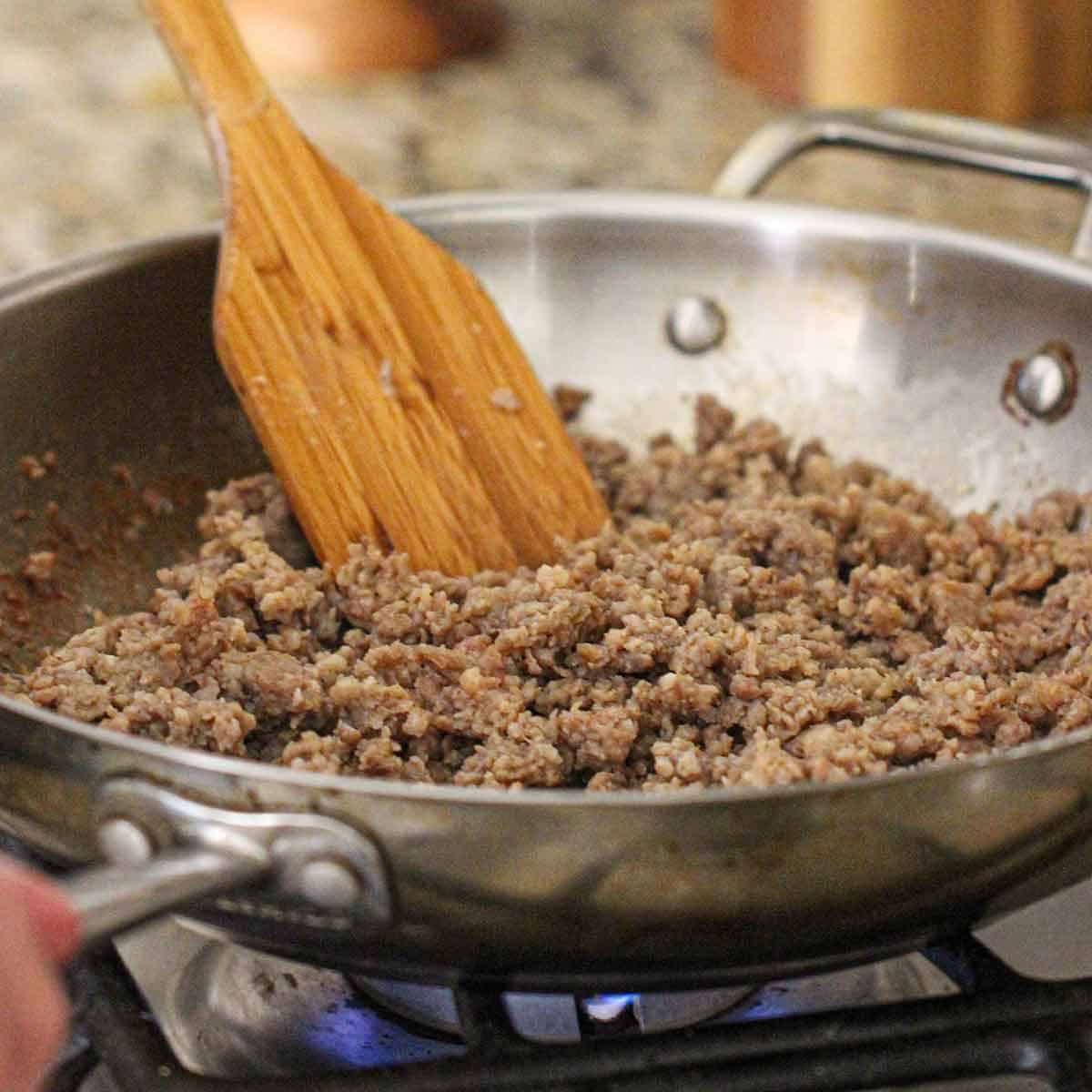 A person using a wooden spatula to stir breakfast sausage while it is being cooked in a skillet on a gas stove.