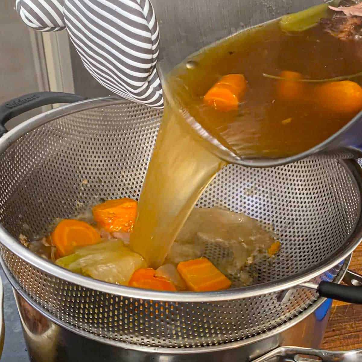 A person transferring roasted turkey stock including roasted vegetables and turkey bones from a large stock pan into a colander over a smaller stock pan.