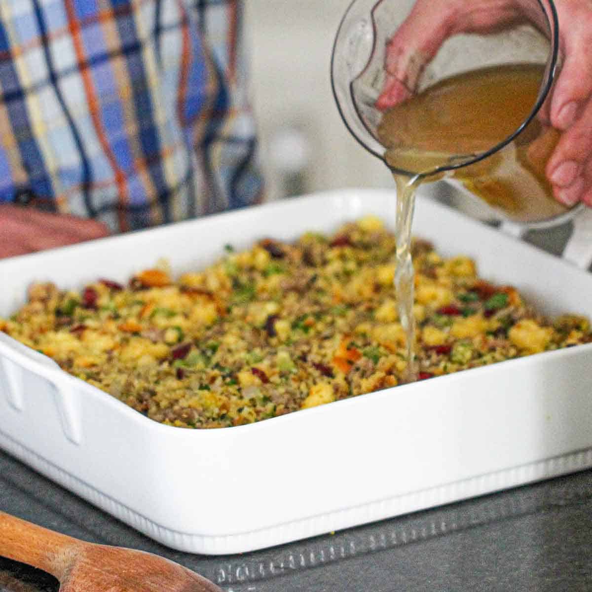 A person pouring chicken stock from a clear measuring cup into a square baking dish that is filled with cornbread dressing.