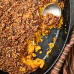 An overhead view of a sweet potato casserole with crunchy pecan topping in an oval baking dish with a portion of the casserole missing and a serving spoon inserted into the dish.