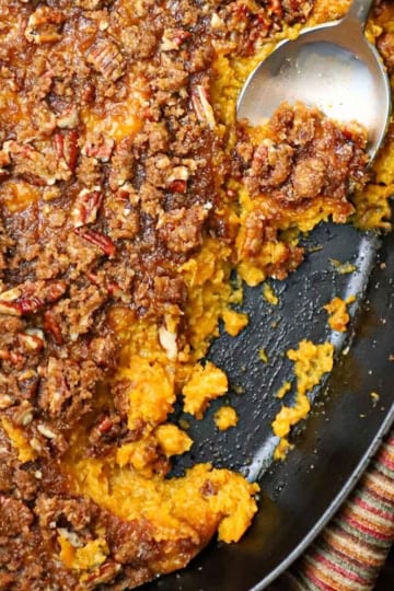 An overhead view of a sweet potato casserole with crunchy pecan topping in an oval baking dish with a portion of the casserole missing and a serving spoon inserted into the dish.
