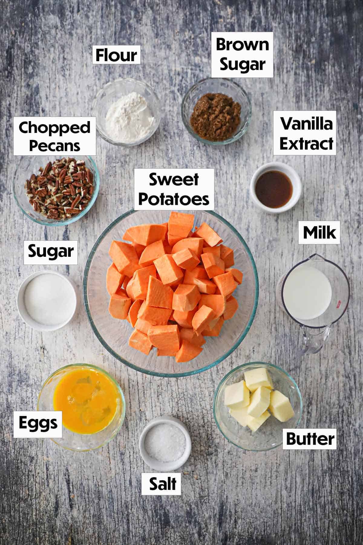 An arrangement of ingredients for sweet potato casserole with crunchy pecan topping on a grey wooden background including cubed sweet potatoes, chopped pecans, flour, sugar, vanilla, milk, eggs, butter, and salt.