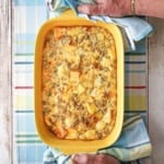 A yellow rectangular baking dish filled with a classic breakfast casserole with the handles of the dish being held by a person holding two linen napkins.