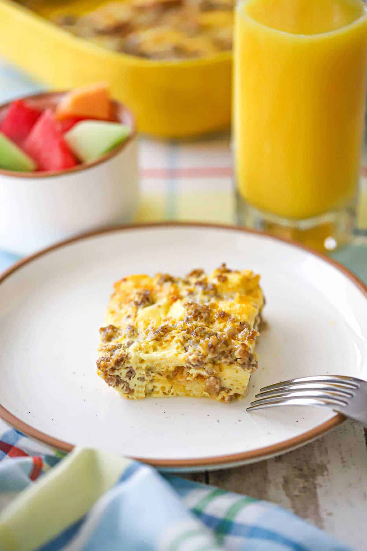 A square piece of classic breakfast casserole on a white plate next to a glass of orange juice and a small bowl of freshly cut fruit.