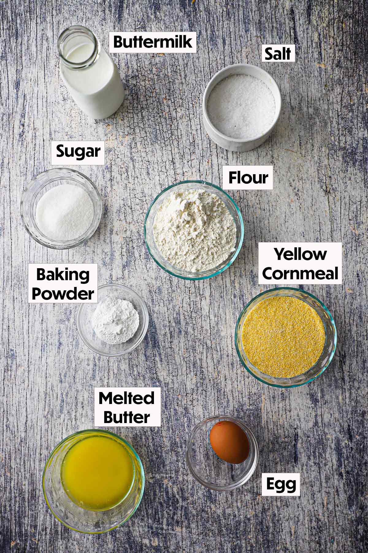 An arrangement of ingredients for homemade cornbread on a grey wooden background including yellow cornmeal, flour, buttermilk, salt, sugar, baking powder, melted butter, and an egg.