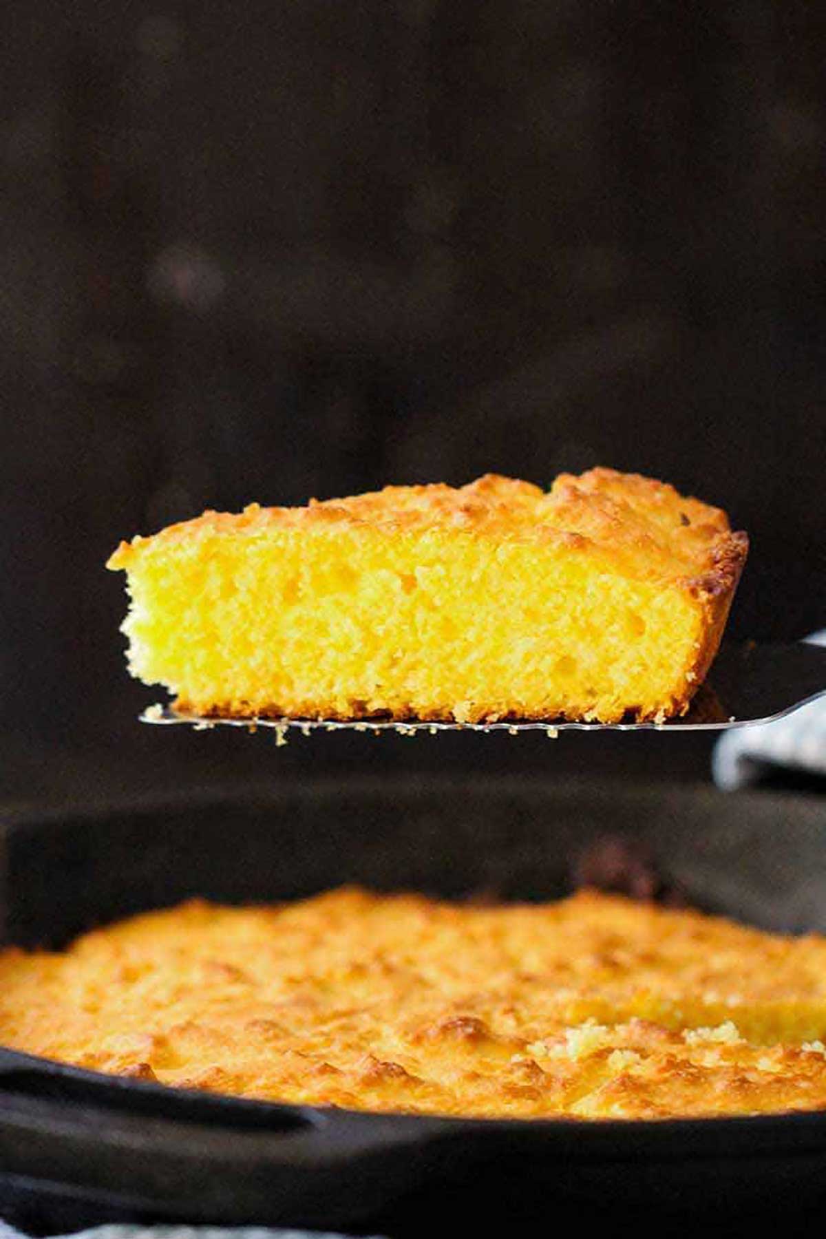 A person using a metal spatula to raised a slice of homemade cornbread from a cast-iron skillet filled with freshly baked cornbread.