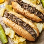 A close-up view of a homemade Philly cheesesteak with another cheesesteak sitting next to it, both on a wrinkled brown piece of sandwich paper.