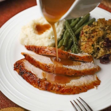 A person pouring turkey gravy from a white gravy boat over a plate filled with three slices of maple glazed whole turkey breast next to cornbread dressing, green beans, and mashed potatoes with gravy.