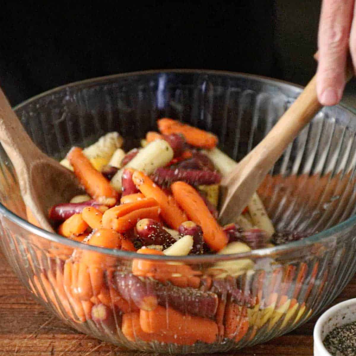 A person using two wooden spoons to toss colorful baby carrots with olive oil, salt, and pepper in a large glass bowl.