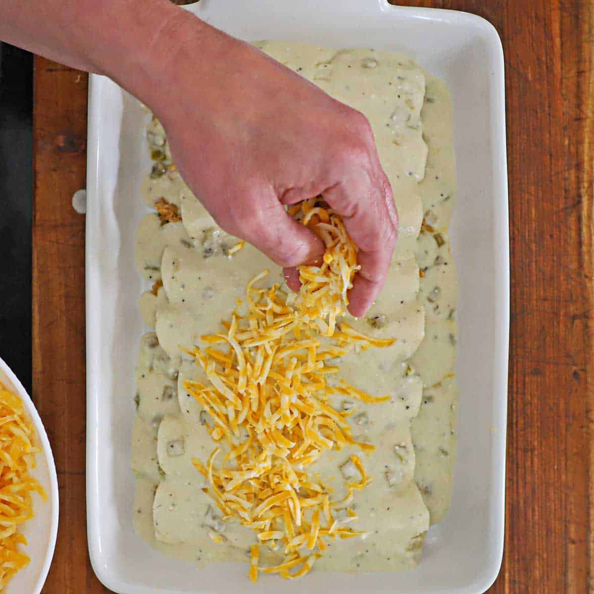 A person adding handfuls of shredded Colby Jack cheese over the tops of uncooked chicken enchiladas with sour cream sauce in a white casserole dish.
