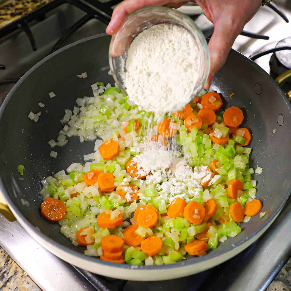 A person dumping all-purpose flour from a small glass bowl into a skillet filled with sautéed carrots, celery, and onion.