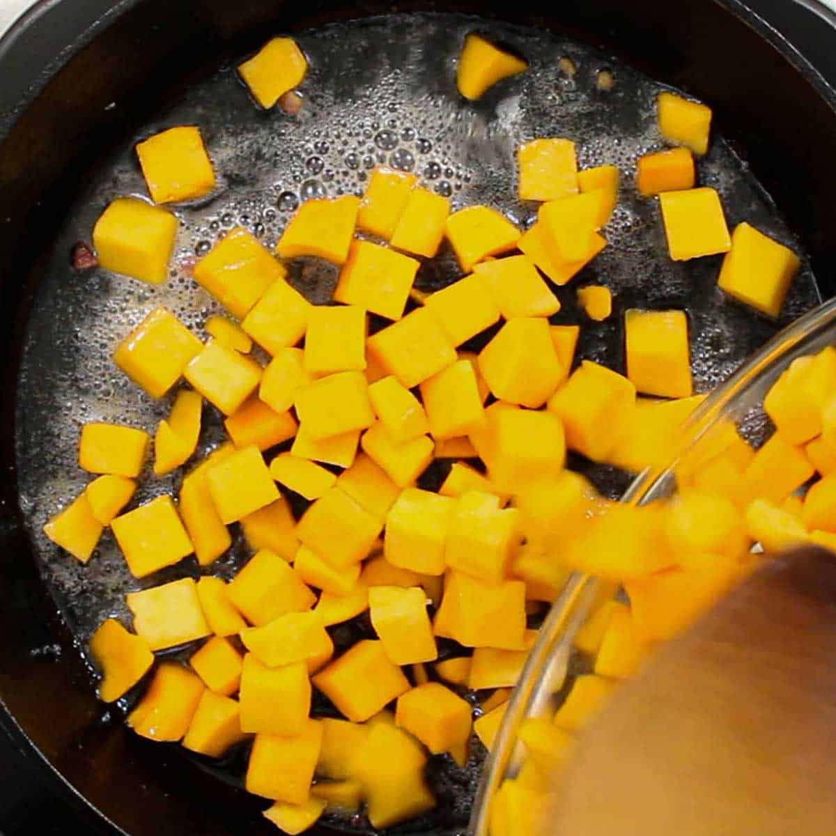A person transferring cubed butternut squash from a large glass bowl into a cast-iron skillet with simmering oil in it.