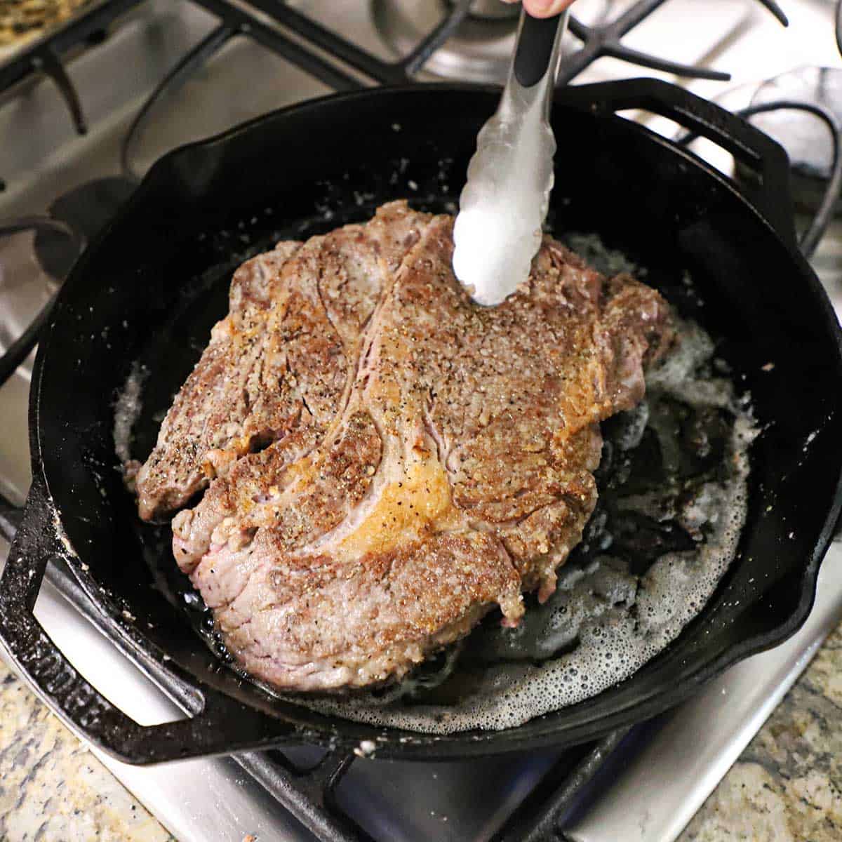 A person using metal tongs to flip are large chuck roast that is being seared in olive oil in a large black cast-iron skillet.