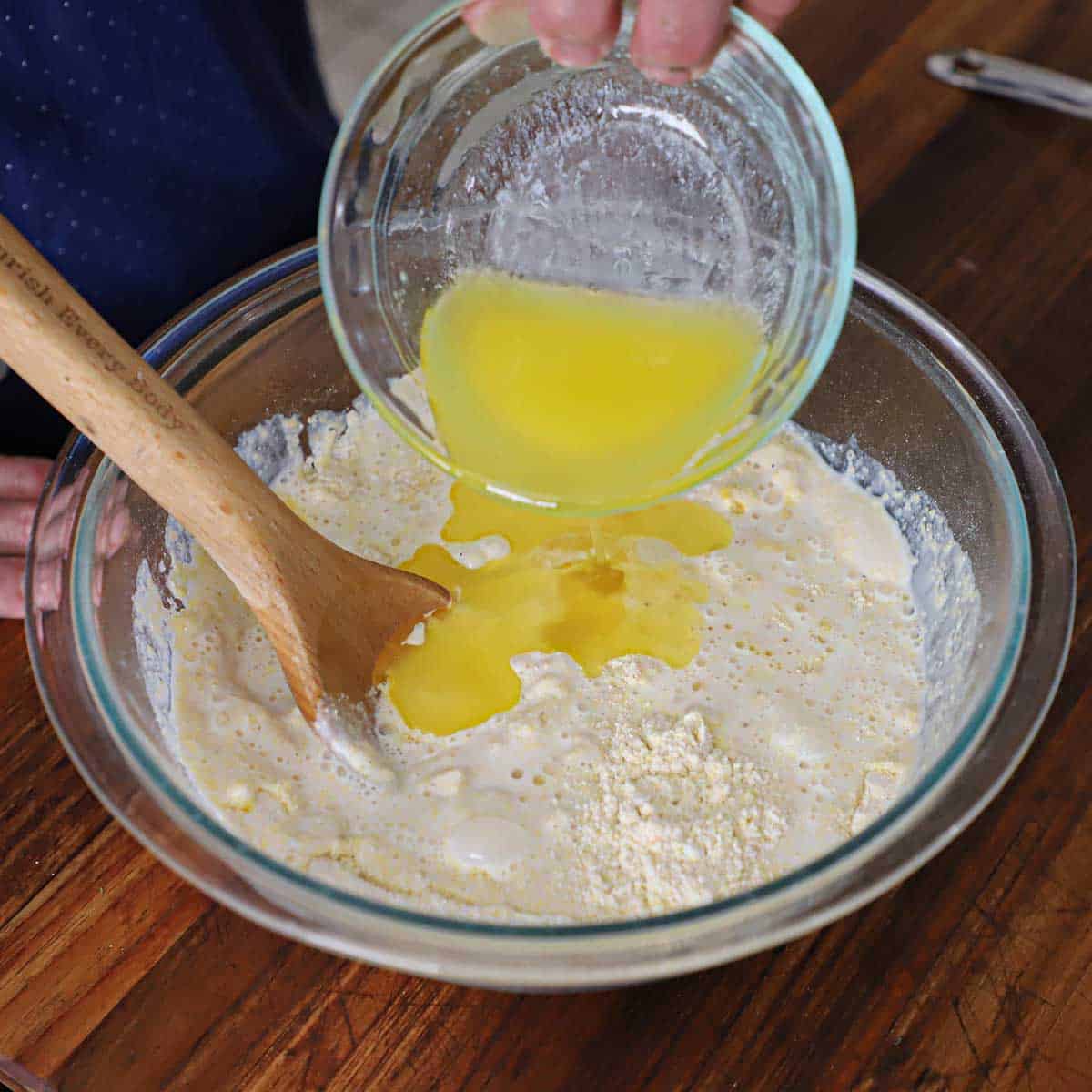 A person pouring melted butter from a small glass bowl into a larger glass bowl filled with dry cornbread ingredients as well as buttermilk and a lightly beaten egg.