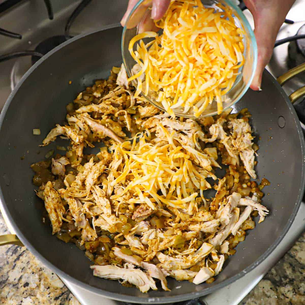 A person transferring shredded Colby Jack cheese from a small glass bowl into a skillet that is filled with chicken enchilada meat.