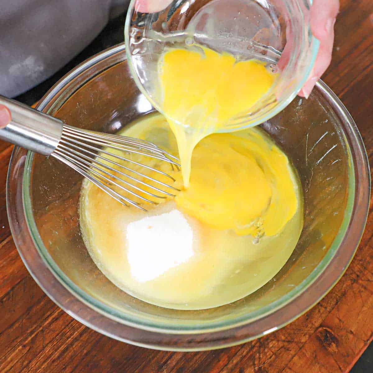 A person pouring lightly beaten eggs from a small glass bowl into another larger glass bowl filled with sugar, corn syrup, and melted butter.