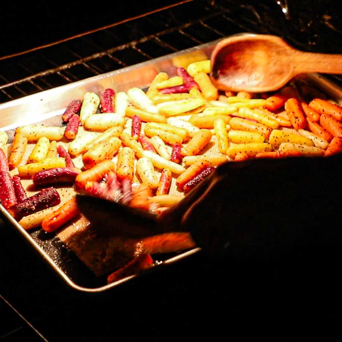 A person using two wooden spoons to toss baby carrots that are being roasted in a large baking sheet pan inside an oven.