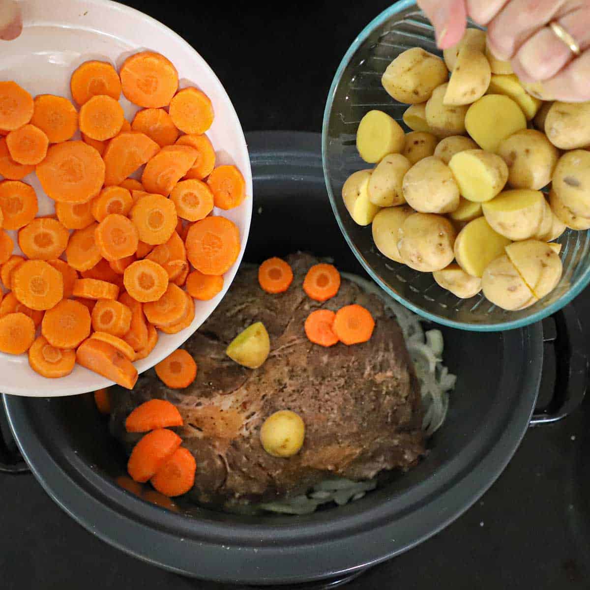 A person transferring sliced carrots from a white bowl with hand and transferring sliced baby potatoes from a glass bowl with his other hand into a slow cooker containing a seared chuck pot roast.