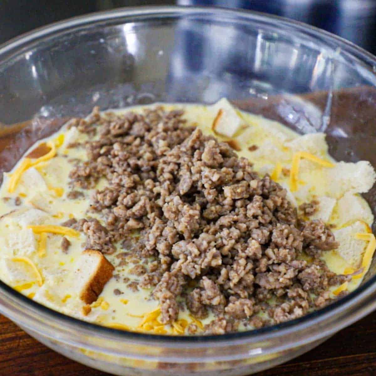 A glass bowl filled with a milk and egg mixture and topped with cooked breakfast sausage, cubed white bread, and shredded cheddar cheese.