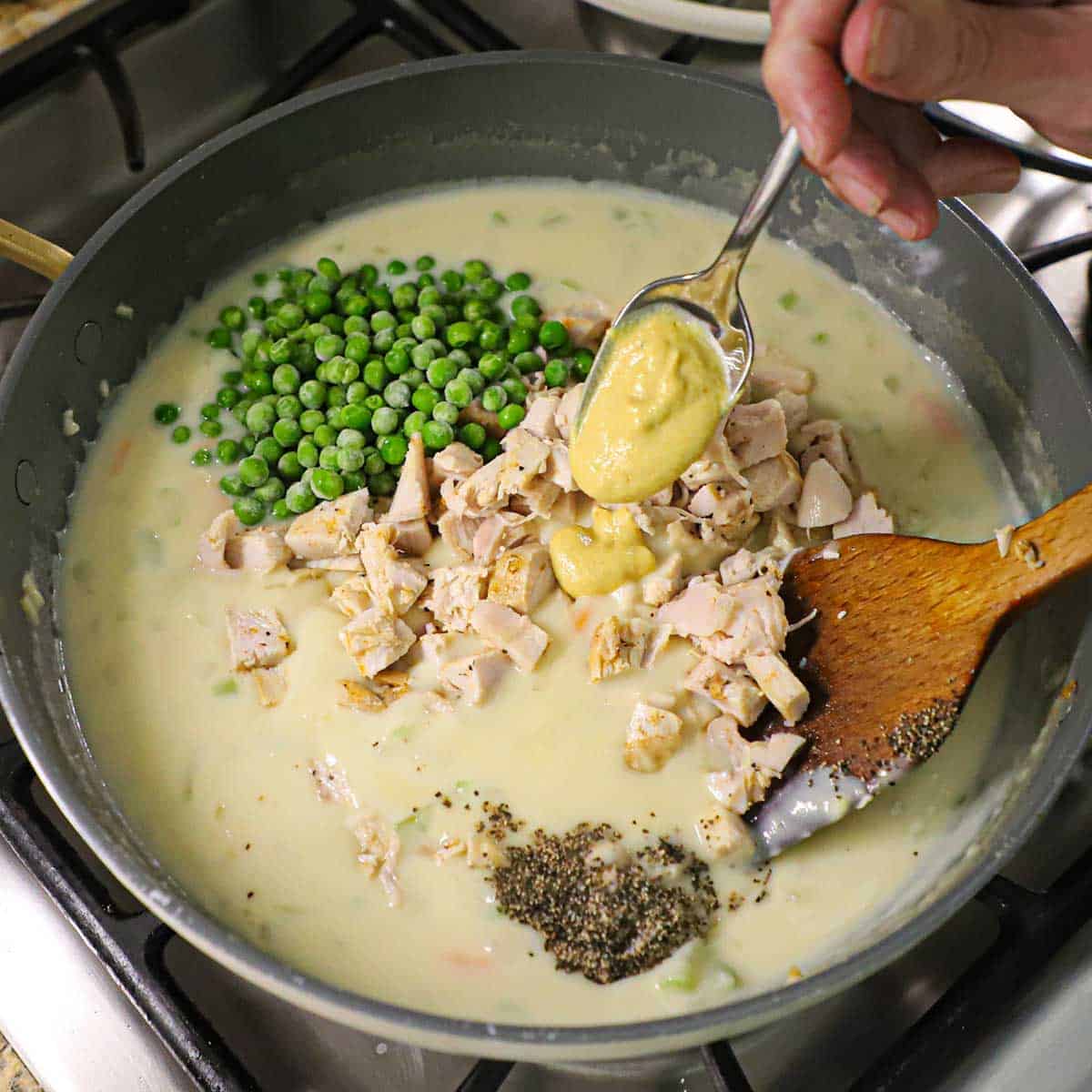 A person adding a spoonful of Dijon mustard into a skillet filled with a thickened pot pie sauce along with sweet peas, cubed leftover turkey, and seasonings.