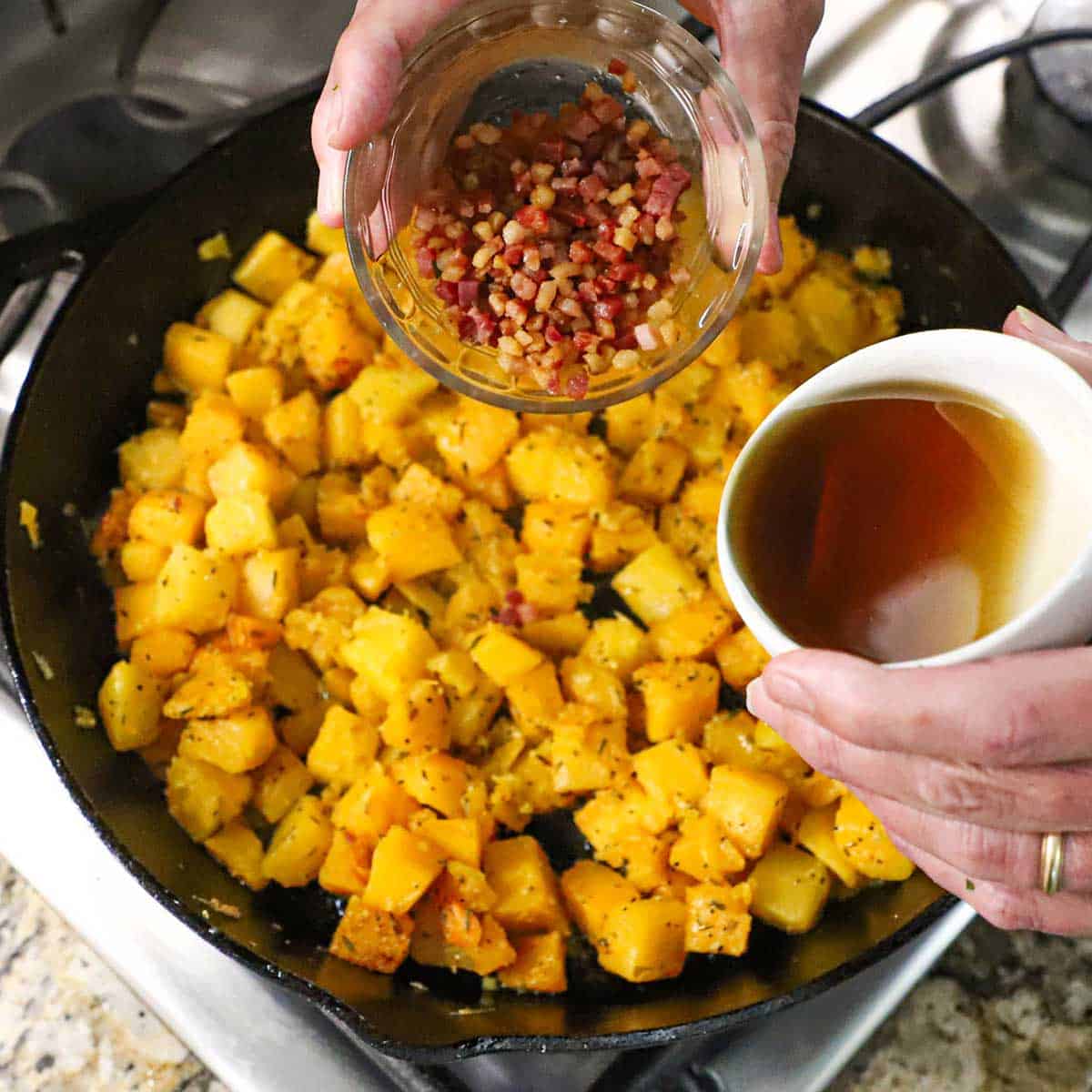 A person holding a small glass bowl of crispy, cooked pancetta in one hand and the other hand holding a small bowl of maple syrup, all over a skillet filled with sauteed butternut squash.