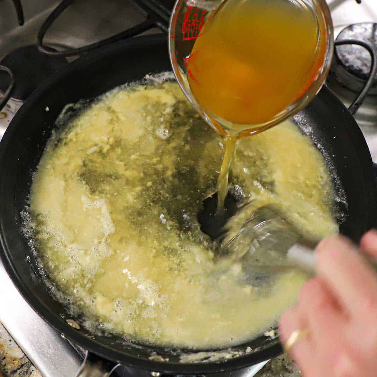 A person using a small whisk to incorporate chicken broth being poured from a glass measuring cup into a skillet filled with a roux all on a gas stove.