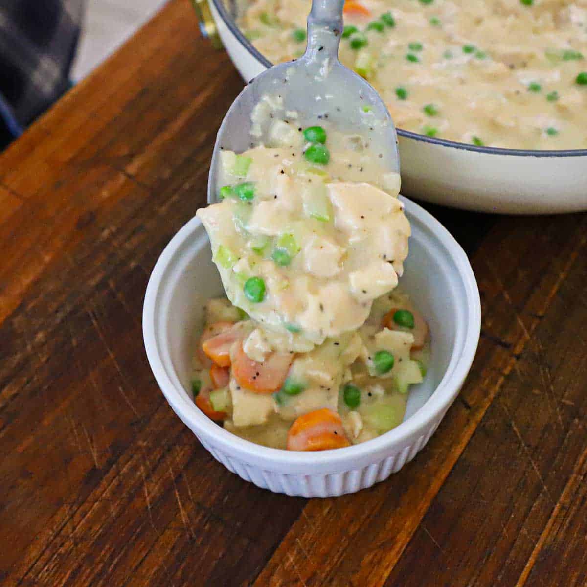 A person using a large spoon to transfer a leftover turkey pot pie filling from a large skillet into a white ramekin.