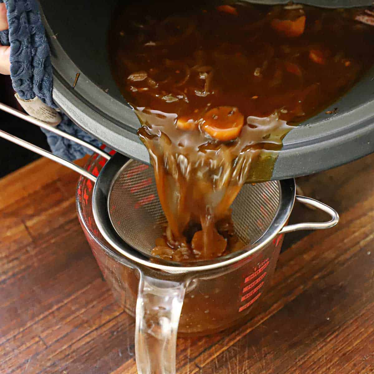 A person straining broth from a slow cooker through a sieve into a large glass measuring cup on a wooden cutting board.