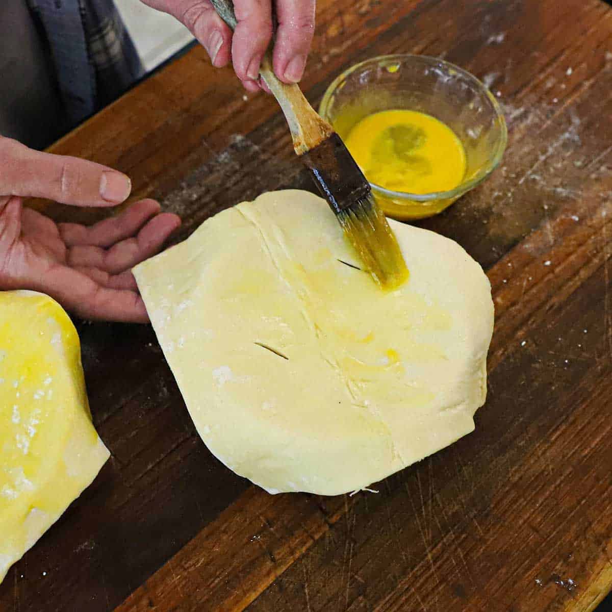 A person using a pastry brush to apply an egg wash over the the surface of a puff pastry sheet that is resting on top of a ramekin filled with leftover turkey pot pie filling.
