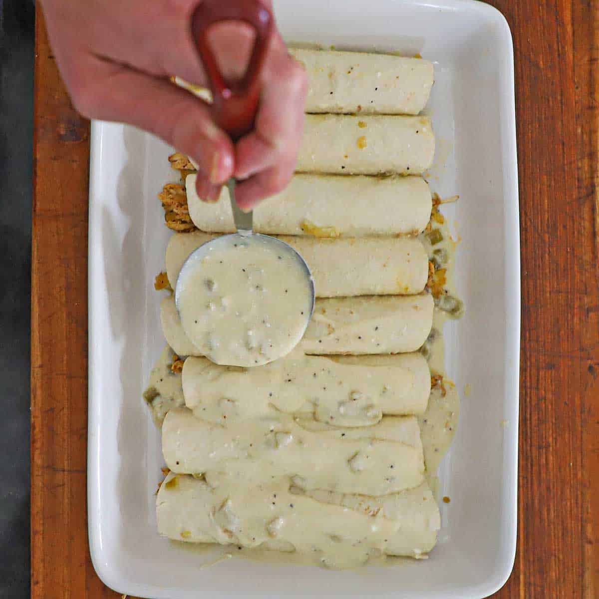 A person using a small ladle to pour a sour cream sauce over the tops of roll chicken enchiladas in a white casserole dish.