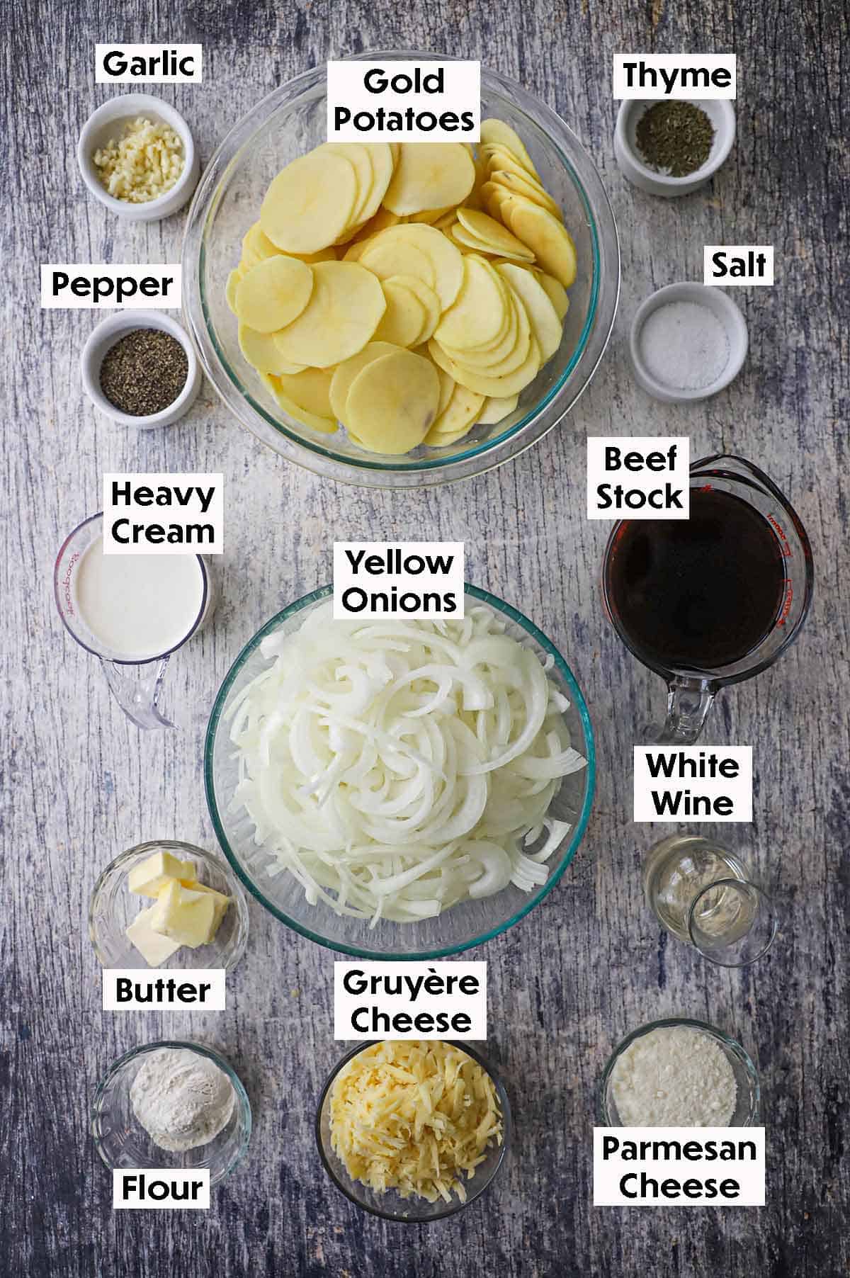 An arrangement of ingredients for French onion au gratin potatoes on a grey wooden background including sliced onions, sliced potatoes, minced garlic, cream, beef stock, wine, butter, cheese, flour, and seasonings.