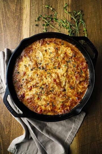An overhead view of a large cast-iron skillet filled with French onion au gratin potatoes with fresh herbs and a grey napkin near the dish.