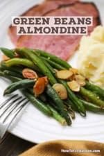 A white dinner plate filled with a serving of green beans almondine next to several slices of smoked ham and a pile of mashed potatoes.