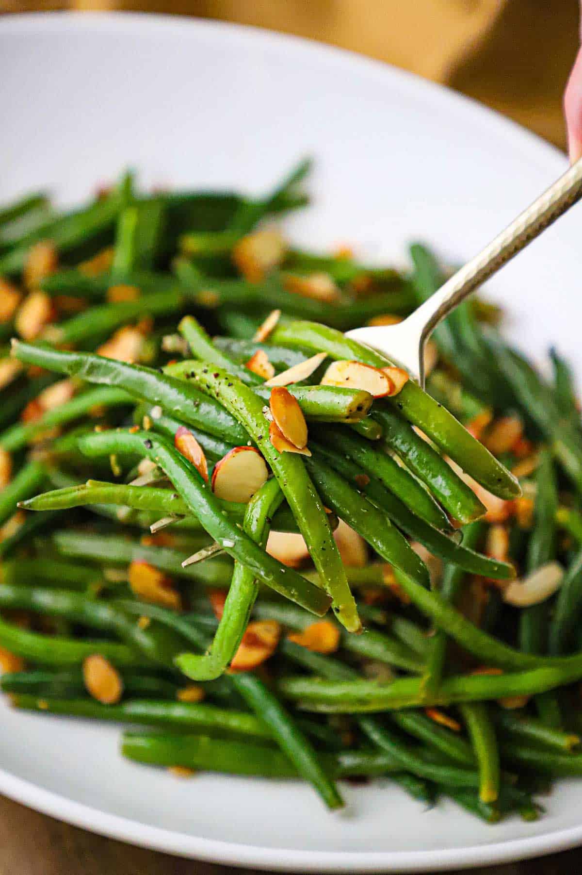 A person using a gold serving utensil to lift a serving of green beans almondine from a white platter of the dish.