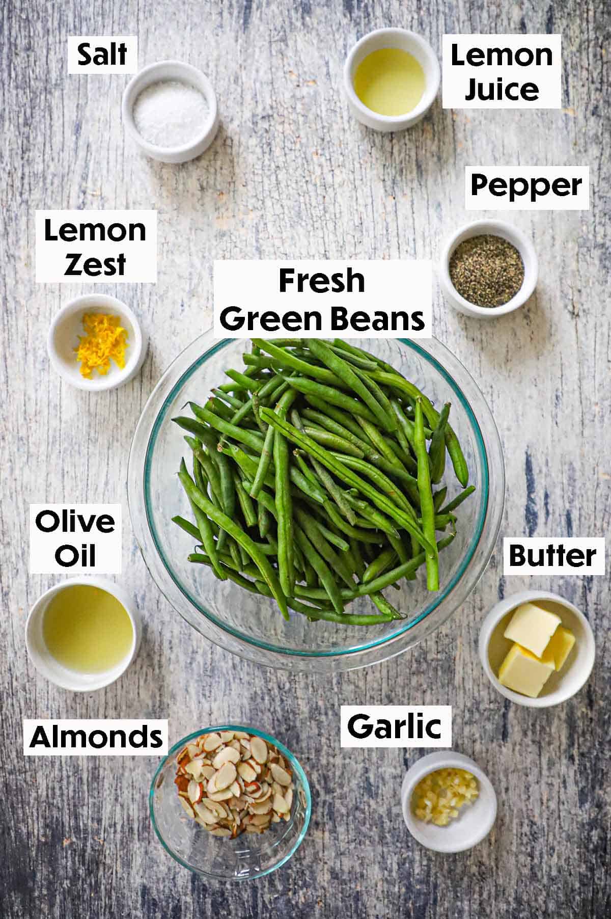 An arrangement of ingredients for green beans almondine on a grey wooden background including a glass bowl of fresh green beans, lemon zest, lemon juice, butter, almonds, garlic, olive oil, and seasonings.