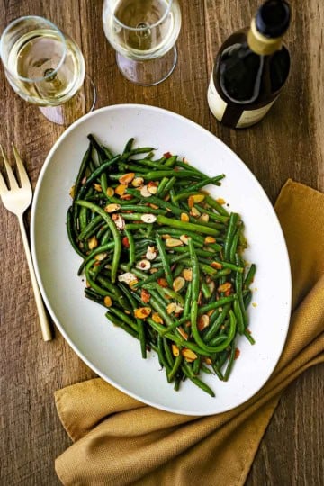 An overhead view of a large oval white platter holding green beans almondine with a bottle of wine and two glass of white wine nearby.