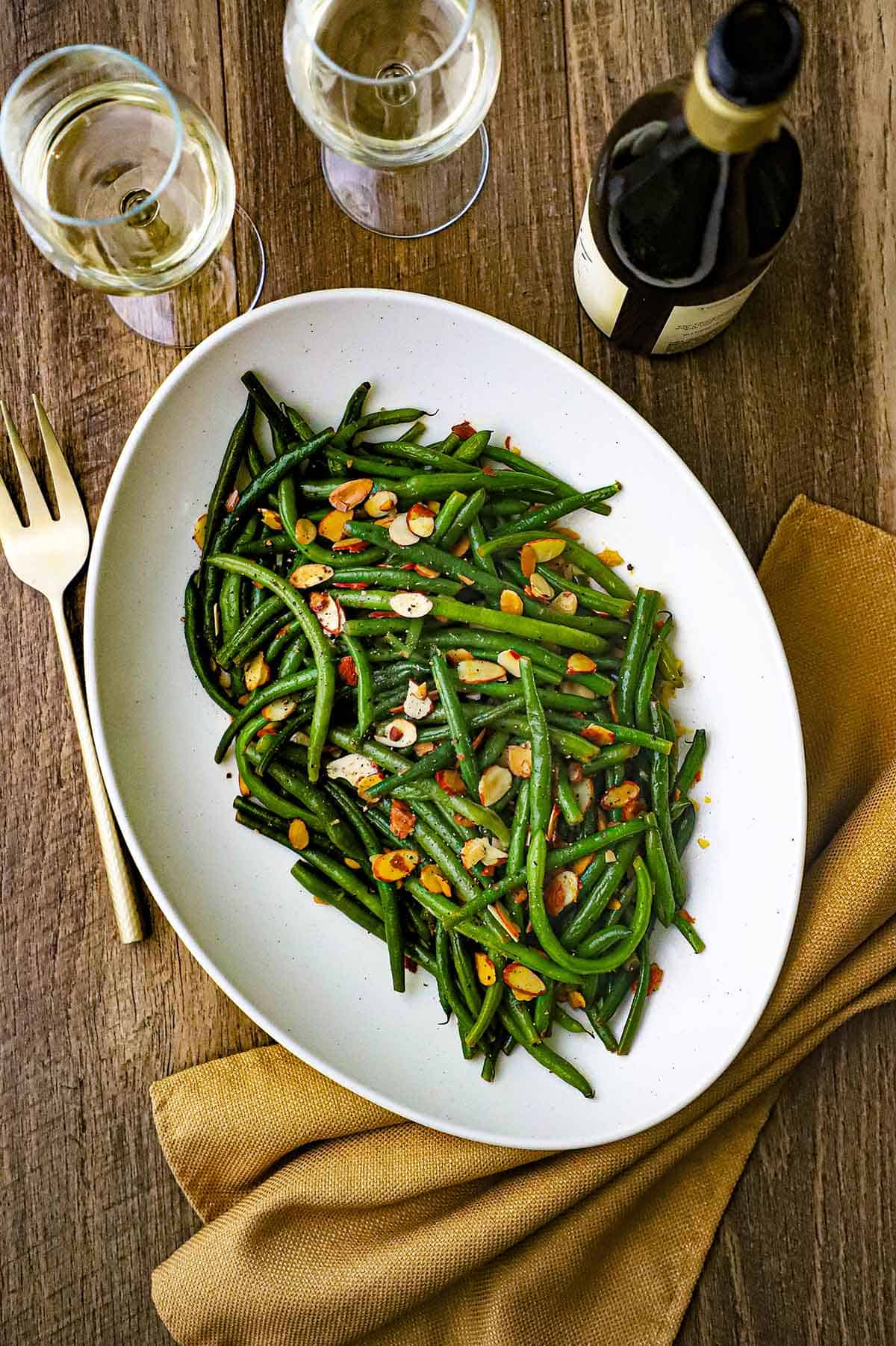 An overhead view of a large oval white platter holding green beans almondine with a bottle of wine and two glass of white wine nearby.
